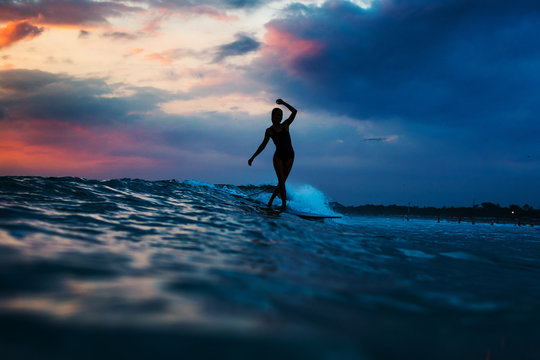 Silhouette of woman in the ocean surfing wave at sunset. Beautiful clouds. Vivid colors.