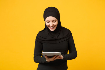 Young arabian muslim employee woman in hijab black clothes hold and work tablet pc computer isolated on yellow wall background, studio portrait. People religious lifestyle concept. Mock up copy space.