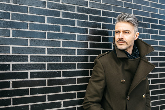 Fashionable businessman with bristle and grey hair looking away at black brick wall.
