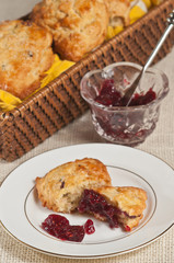 top view, close distance of a cranberry, ginger, cream scone, torn open and covered with raspberry jam on a round, white dish, with a crystal bowl  of jam and a wicker basket of scones in background