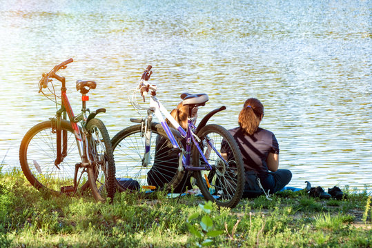 Two girls sitting on the bank of the river at two bicycles having rest and talking. Girls friendship.