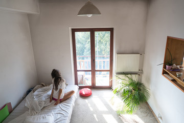 Young woman sitting on bed and looking through the window.
