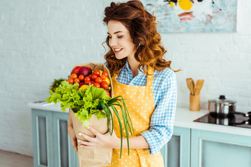 smiling woman in polka dot apron holding paper bag with ripe fruits and vegetables