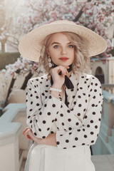 Outdoor fashion portrait of young beautiful lady wearing stylish straw wide-brimmed hat, polka dot blouse, trendy shell earrings, posing in street © Victoria Fox