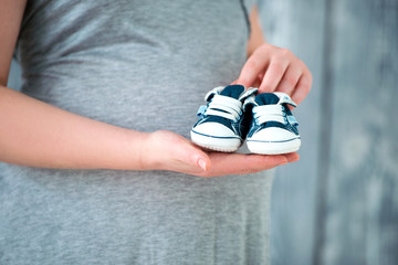Happy mother in anticipation of the birth of her son. Pregnant woman holding baby booties sneakers on tummy background. Close-up of a pregnant belly.