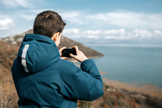 A boy by the lake is taking a picture with phone camera