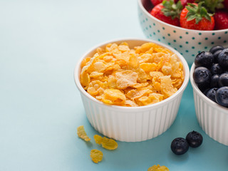Concept of healthy gluten free breakfast with berries and corn flakes in the white bowls on a blue background, three plates, top view