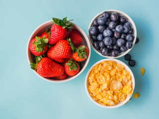 Concept of healthy gluten free breakfast with berries and corn flakes in the white bowls on a blue background, three plates, top view