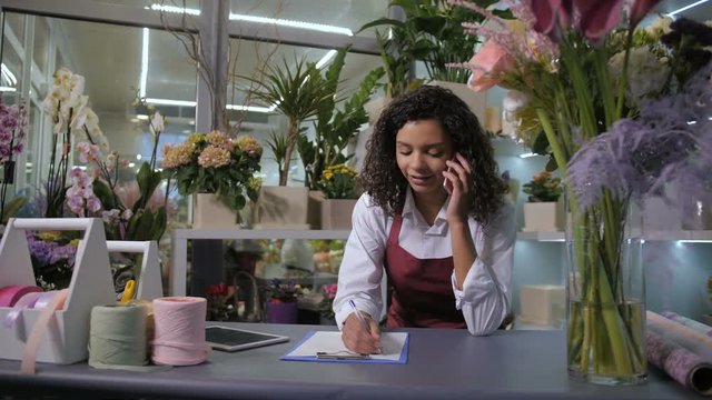 Pretty mixed race florist communicating on cellphone with important client taking big order for flower decoration of wedding hall. Curly woman noting at workplace surrounded by plants in flower shop