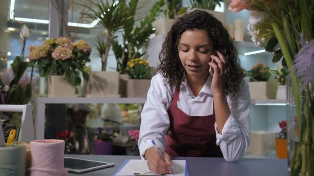Attractive female florist conversating on smartphone with customer taking order for Valentine's day flower arrangement. Smiling mixed race woman working in flower shop surrounded by flowering plants