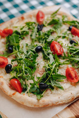 Neapolitan pizza with tuna, cheese, arugula, basil, tomatoes, olives, sprinkled with cheese on a wooden table on a tablecloth in a cell with a place for the text.