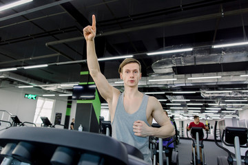 Fototapeta na wymiar Young happy handsome man with hand up in fitness club. Cardio workout, running on treadmill. Healthy lifestyle, guy and woman training in gym.