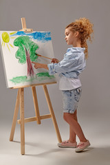 Charming school girl is painting with a watercolor brush on an easel, standing on a gray background.