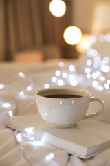 Cup of coffee and book on bed against blurred background. Space for text
