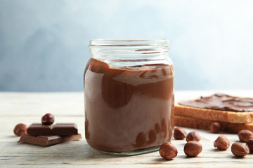 Glass jar with tasty chocolate cream and hazelnuts on wooden table against color background