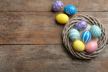 Wicker nest with painted Easter eggs on wooden table, top view. Space for text