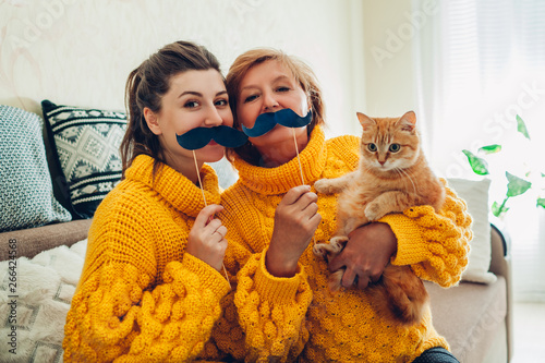 Senior mother and her adult daughter taking selfie with cat using photo booth props at home. Mother's day concept.