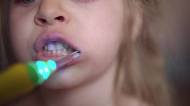 Close-up of cute blonde child brushing her teeth.Toothbrush glows with colored lights, helping kid to see every baby tooth in mouth. Concept of oral hygiene and healthy lifestyle.