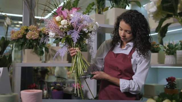 Professional female florist completing gorgeous composition of floral bouquet cutting flower stems with pruner in floristical salon. Attractive mixed race woman in apron creating flower arrangement