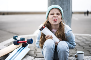 A naughty girl in a blue sweater and hat was riding a longboard and sat down on the curb to drink...
