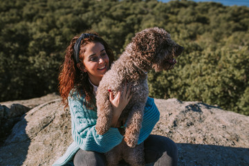 A young beautiful woman is sitting with her dog on the stone. She is hugging her pet while they are looking the landscape. They have brunette curly hair.