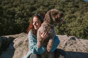 A young beautiful woman is sitting with her dog on the stone. She is hugging her pet while they are looking the landscape. They have brunette curly hair.