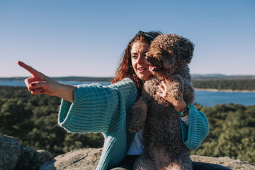 A young brunette woman is pointing at a place in the middle of the nature with her hand while she is holding her dog with the other arm. She is wearing a blue jacket and black jeans.
