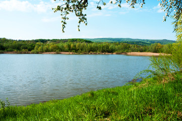 Spring panorama of a forest lake against a background of blue sky and distant mountains