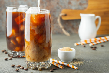 Ice coffee in a tall glass with cream poured over and coffee beans. Cold summer drink on a brown rusty background