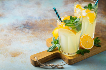 Two glass with lemonade or mojito cocktail with lemon and mint, cold refreshing drink or beverage with ice on rustic blue background. Copy space - obrazy, fototapety, plakaty