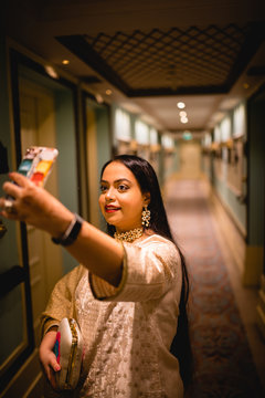 waist up picture of a girl wearing traditional wear, ear ring, necklace standing in a vintage corridor of with lamps, decor and carpet floor.Taking a picture of self.