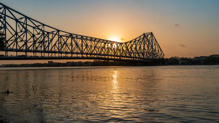 Silhouette of Howrah Bridge at the time of Sunrise.  Howrah Bridge is a bridge with a suspended...
