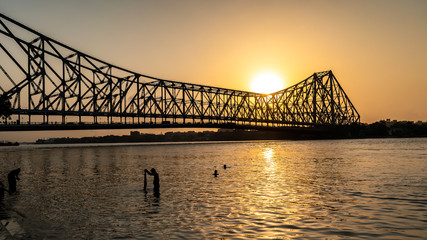 Fototapeta na wymiar Silhouette of Howrah Bridge at the time of Sunrise. People bathing and making rituals in the River Ganges.