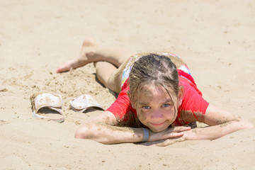 Dirty girl in the sand is lying on beach in red tshirt in hot sun.