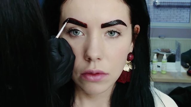 Woman cosmetologist hand apply paint with brush on the eyebrows of a beautiful patient. Eyebrow correction procedure close-up. Professional care for face in beauty saloon