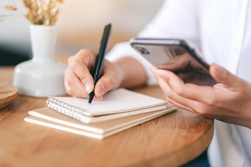Closeup image of a woman using mobile phone while writing on a blank notebook on wooden table