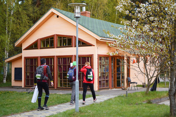 Fototapeta na wymiar Three athletes wearing running backpacks man and two women participants of orienteering or rogaining sport competition walking on pathway to wooden house information centre.