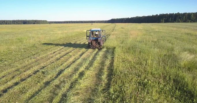 drone follows tractor with big wheels and mower cutting off grass and leaving trace against beautiful landscape