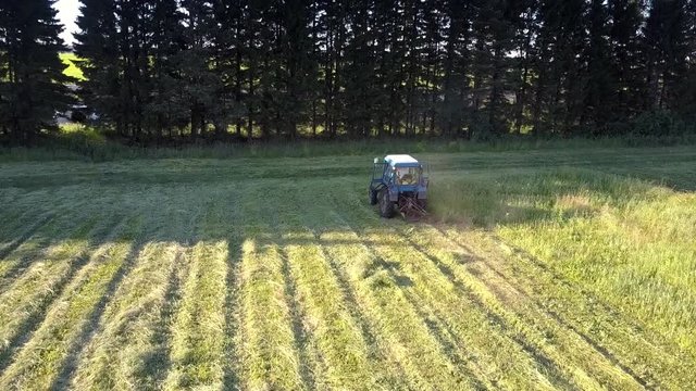 close flight over tractor mowing thick green hay grass to swathes in trees shadow on hot summer day