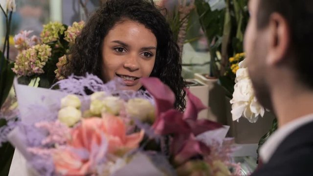 Closeup of happy female florist giving gorgeous floral arrangement with fresh exotic flowers and decorative herbs to satisfied customer. Smiling mixed race woman enjoying beauty of own floral creation