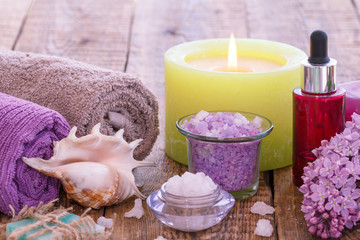 Soap, burning candle, bowls with sea salt, bottle with aromatic oil, lilac flowers and towels on wooden background.