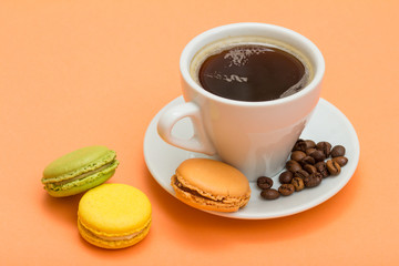 Cup of coffee with coffee beans and delicious macarons cakes of different color on peach background.
