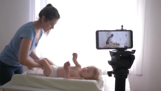 modern blogger work, young mother blogger changes nappy to small baby boy while recording training video for subscribers live on mobile phone at home