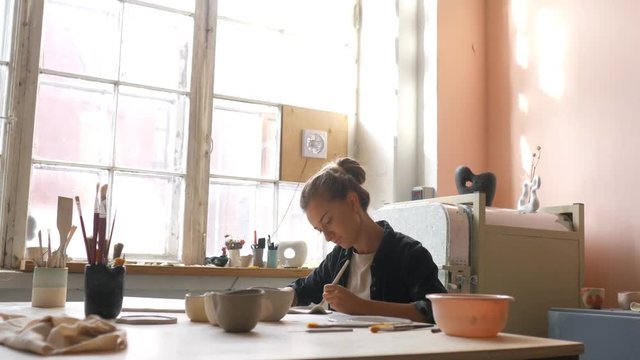 woman artist paints on ceramics in a workshop at a large bright window