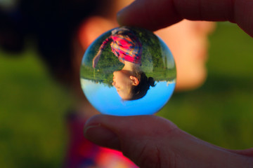 Blurred Portrait Of A Young Girl Reflection In A Glass Ball Outdoors. Abstract People. People, Travel Concept. Female Fingers Holding Glass Ball. 