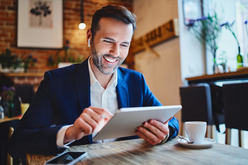 Businessman sitting in cafe looking at tablet during phone call through wireless headphones - Powered by Adobe