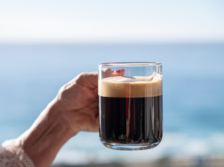 Hand holding a glass of coffee by the ocean