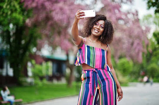 Fashionable african american woman in pink striped jumpsuit posed at spring bloom street and making selfie by mobile phone. - Powered by Adobe