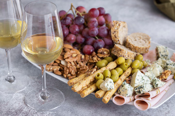 Two glasses of white wine and plate with different snacks. Blue cheese, olives, baguette slices, grissini, ham, grapes and nuts. Wine snacks set background.