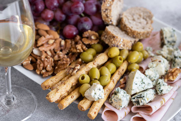 Glass of white wine and plate with different snacks. Blue cheese, olives, baguette slices, grissini, ham, grapes and nuts. Wine snacks set background.